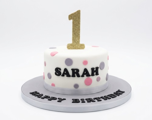 White round birthday cake with pink and grey polka dots, a gold glitter number one topper, and a personalised name.