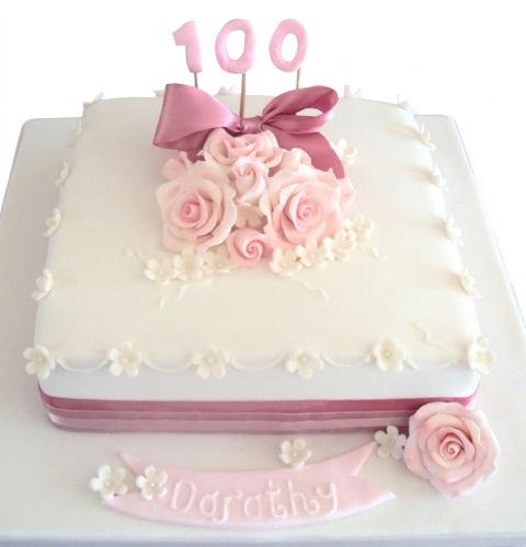 Elegant white square cake with pink sugar roses, bow, and '100' custom age topper.
