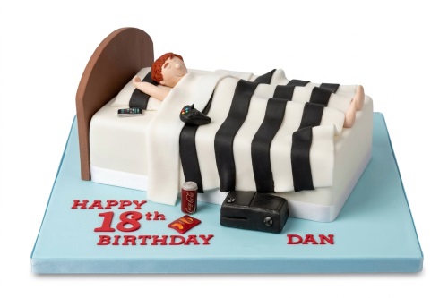 Novelty birthday cake featuring a sleeping figure in bed with a gaming controller, console, and snacks, on a blue board with custom age message.