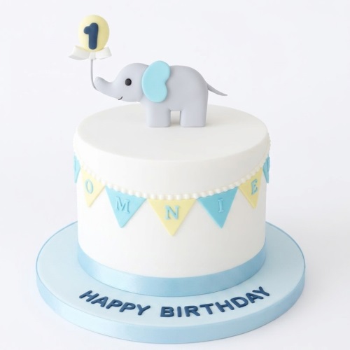 White single-tier cake with a grey elephant topper holding a yellow balloon with the number '1', blue and yellow bunting, and 'Happy Birthday' on the cake board.