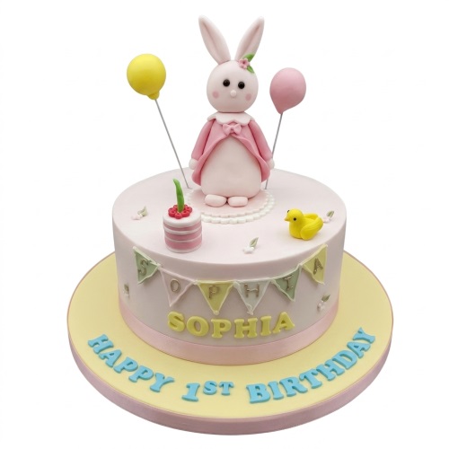 Pink round cake with a bunny figure, balloons, and a duck, with personalised message.