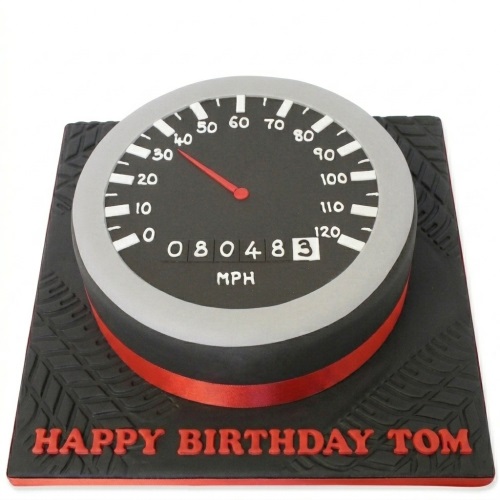 Round black speedometer cake with red needle and mileage, on a black board with tire treads and red ribbon.