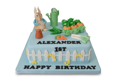 Peter Rabbit themed birthday cake with garden vegetables, watering can, and picket fence. Features a custom age and name.