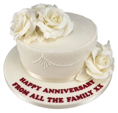 Elegant two-tier white anniversary cake with white sugar roses, delicate lace piping, and a custom message.