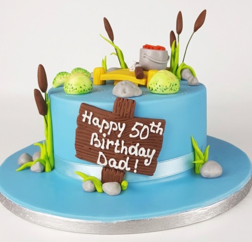 Blue fishing theme birthday cake with cattails, lily pads, fishing bucket, and a custom message sign.