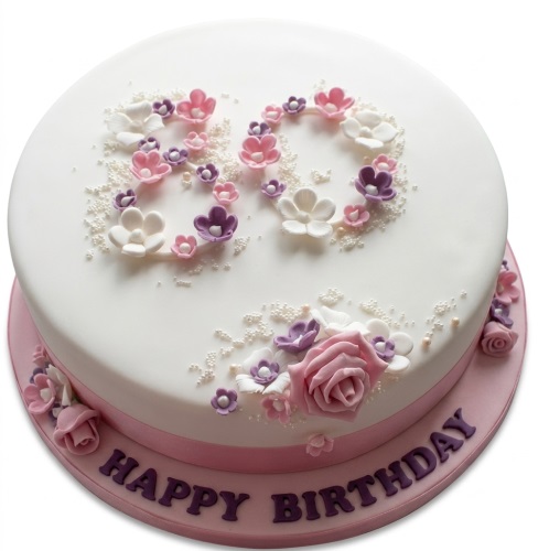 Round white birthday cake with pink, purple, and white sugar flowers, edible pearls, and a pink ribbon.