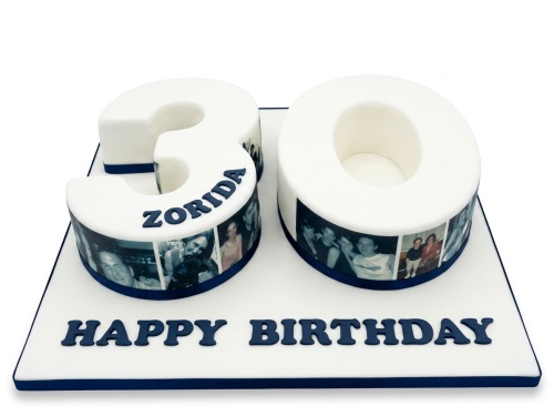 White number cake in the shape of '30' with a photo strip around the base and a custom name on top. The cake board says 'Happy Birthday'.