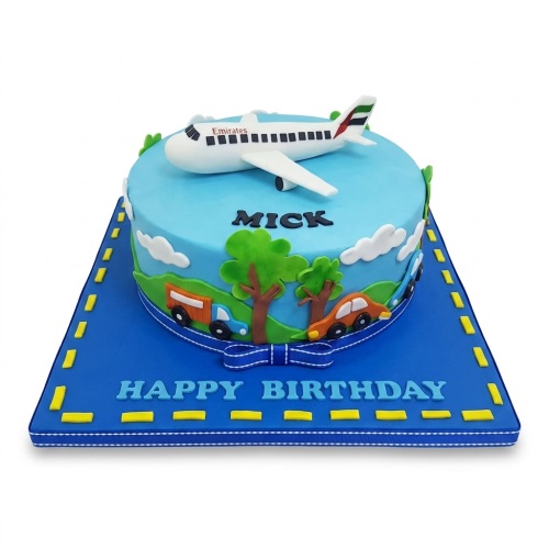 Round blue birthday cake with a white aeroplane topper, cars, trees, and clouds. Features a custom name and 'Happy Birthday' message.