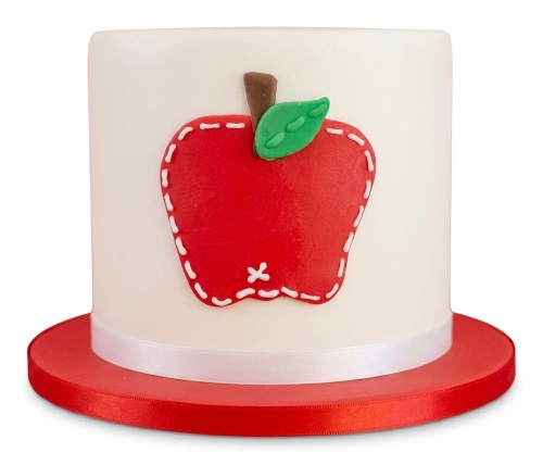 White round cake with a red apple decoration, green leaf, brown stem, and white ribbon on a red board.