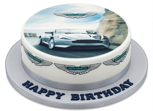 Round white birthday cake with an edible image of a silver Aston Martin car on top and Aston Martin logos on the side, with 'Happy Birthday' written on the board.
