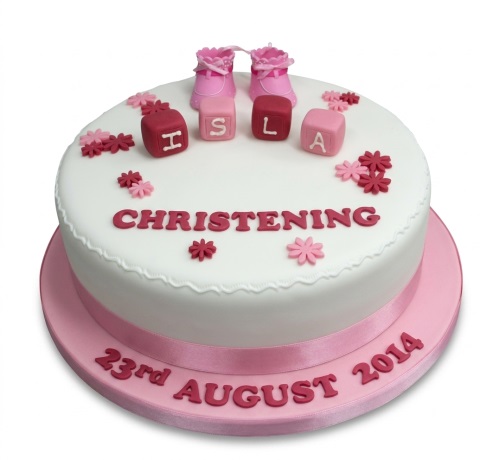 White christening cake with pink baby booties, building blocks, and small flowers, on a pink cake board.