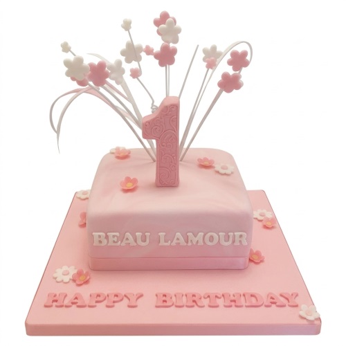 Two-tiered pink square cake with a large number 1 topper, white and pink floral spray decorations, and scattered small flowers.