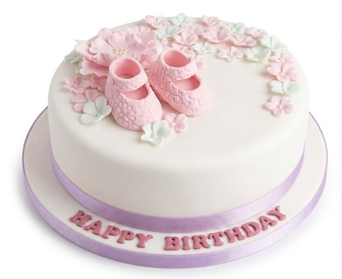 White round cake with pink baby booties, pink and green sugar flowers, and a purple ribbon with 'Happy Birthday' on the board.