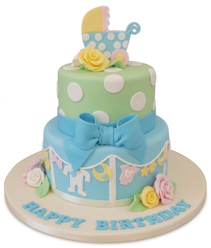 Two-tier baby birthday cake with green polka dot top tier, blue bottom tier with baby bunting, pram topper, and pastel roses.