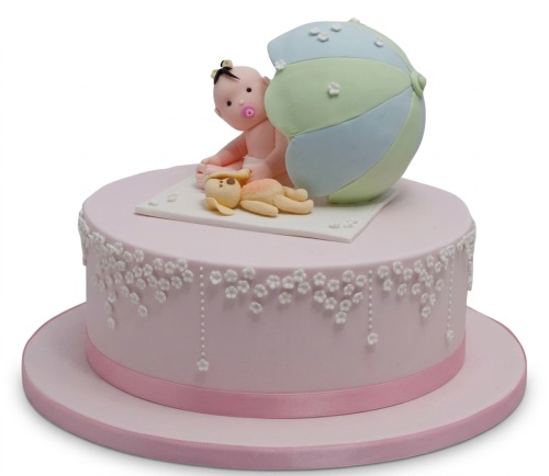 Pink baby shower cake with baby figure, teddy bear, and pastel umbrella decorations, adorned with white flowers.
