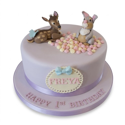 Round lilac cake with fondant deer and rabbit figures, pink and yellow rosettes, and a plaque with a bow.