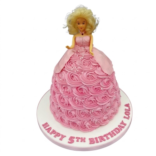 Pink princess doll birthday cake with buttercream rosettes and white sprinkles, on a white cake board.
