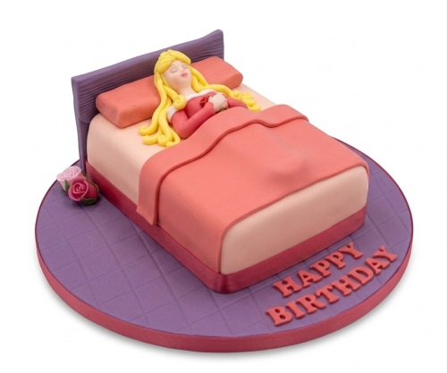 Sleeping princess in a bed shaped birthday cake with pink bedding and purple headboard.
