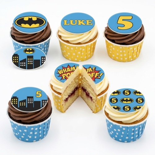 Batman themed cupcakes with blue and yellow toppers featuring the Batman logo, city buildings, and comic book sound effects.