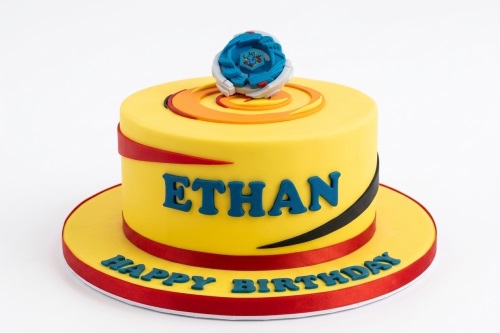 Yellow single-tier birthday cake with a blue Beyblade topper, circular orange and red design, and 'Happy Birthday' message.