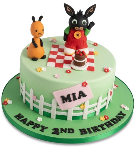 Round light green cake with Bing Bunny and Flop figures, a picnic blanket, small cake, white fence, and colourful flowers.