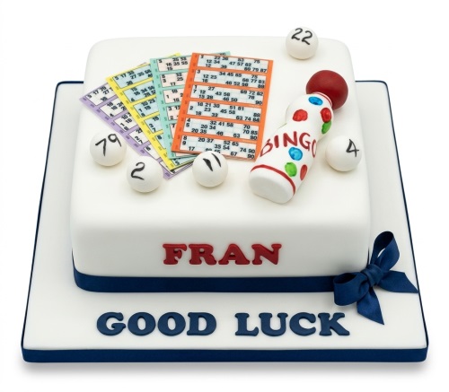 Two-tier square white cake with bingo theme decorations, including bingo cards, shaker, and bingo balls. Features a dark blue ribbon and bow.