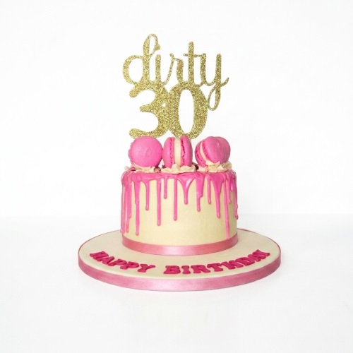 Round cream cake with pink drip, pink macarons, and a gold glitter "Dirty 30" topper.