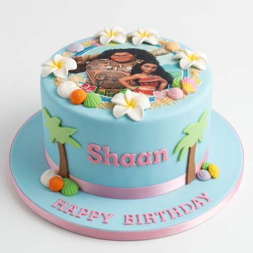 Round blue Moana theme birthday cake with edible image of Moana and Maui, sugar flowers, shells, and palm trees.