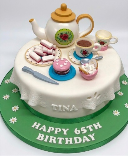 White round cake with edible teapot, teacups, sugar, and biscuits, on a green board with daisies.
