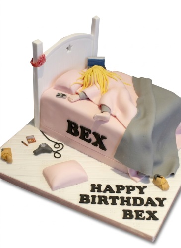 Novelty birthday cake featuring a sleeping figure in a pink bed with a white headboard and custom name.