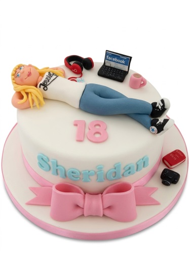 Round white birthday cake with a blonde girl figure, laptop, headphones, camera, and a pink bow.