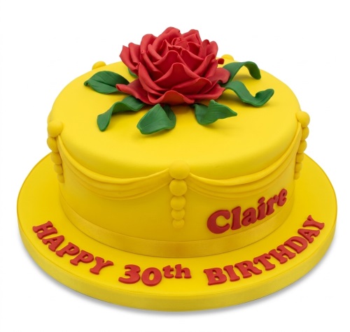Round yellow birthday cake with a large red rose, green leaves, and custom message.