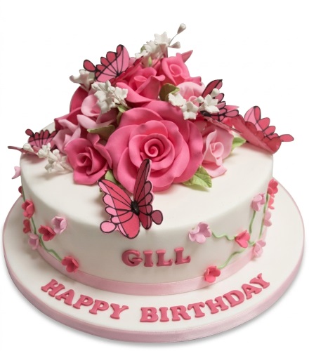 White round birthday cake with pink sugar roses, white flowers, and pink butterflies, with a custom name and 'Happy Birthday' message.