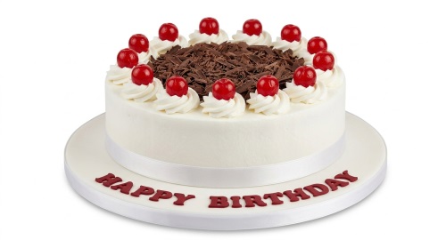 Round white birthday cake with chocolate shavings, red cherries, and cream swirls, with "Happy Birthday" written on the board.