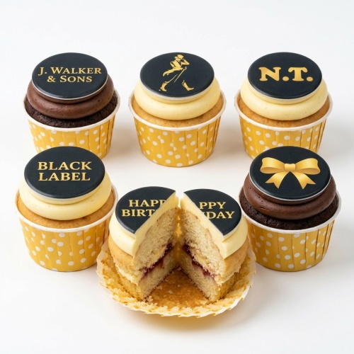 Set of six cupcakes with black and gold whisky themed toppers, including 'J. WALKER & SONS', 'BLACK LABEL', a walking man logo, 'N.T.', a gold bow, and 'HAPPY BIRTHDAY' split across two cupcakes. One cupcake is cut open showing vanilla sponge with jam filling.