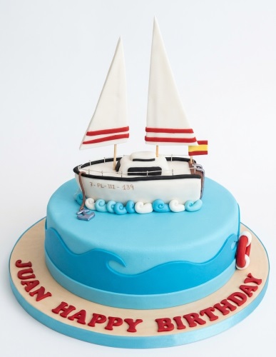 Round blue fondant cake with a 3D sailing boat topper, white sails, red stripes, and a red lifebuoy.