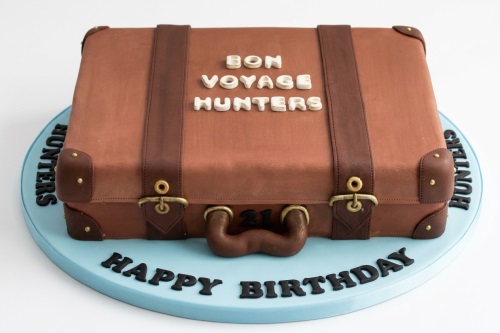 Brown suitcase cake with dark brown straps, gold buckles, and white 'Bon Voyage Hunters' message, on a blue 'Happy Birthday' cake board.