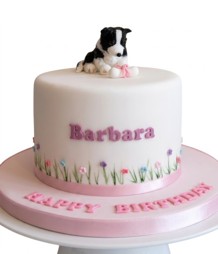 White fondant birthday cake with a black and white dog figurine and a bone, decorated with green grass, small flowers, and a pink ribbon.
