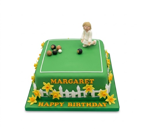 Square green birthday cake with a female bowls player figurine, bowls, white picket fence, and yellow daffodil decorations.