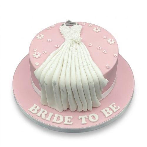 Round pink cake with a white fondant wedding dress, small white and pink flowers, and 'BRIDE TO BE' text on the board.