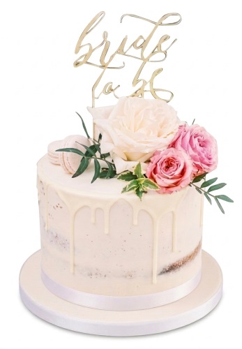 Round semi-naked cake with white drip, pink and cream roses, green leaves, macarons, and a gold 'Bride to Be' topper.