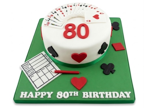 White round 80th birthday cake with playing cards and bridge score sheet on a green board.