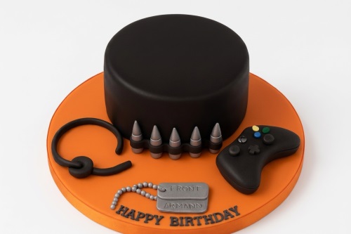 Black gaming theme birthday cake with game controller, bullet belt, and dog tags on an orange board. Personalised message example.
