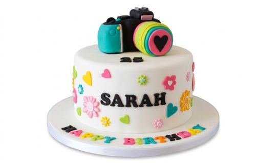 Round white birthday cake with a colourful camera topper, vibrant fondant flowers, hearts, and a custom age number.