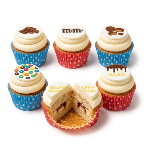 A selection of cupcakes with white frosting and edible toppers featuring chocolate bars, M&M's logo, Reese's Peanut Butter Cups, and scattered M&M's. One cupcake is cut in half revealing a jam filling. Some toppers show 'Happy Birthday' and 'ALEX' text, indicating customisation.