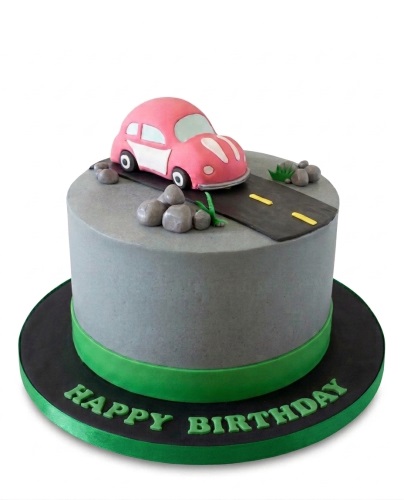 Round grey birthday cake with a pink car, road, rocks, and green 'Happy Birthday' message.