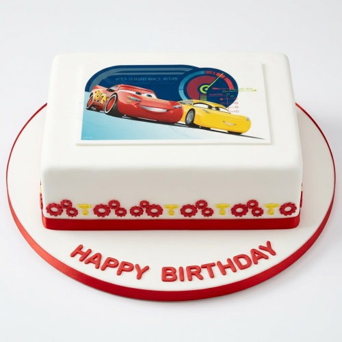 Square white birthday cake with edible Disney Cars image of Lightning McQueen and a yellow car, red and yellow gear decorations, and "Happy Birthday" message.