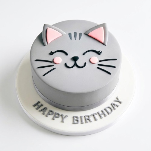 Round grey cat face birthday cake with pink ears and cheeks, black eyes, nose, mouth, and whiskers, on a white board with 'Happy Birthday' message.