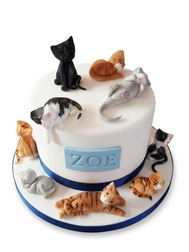 Round white birthday cake with multiple fondant cat figurines and a personalised name plaque.