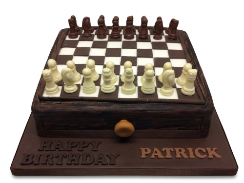 Rectangular brown cake with white and dark chocolate chess pieces on a chessboard design, with 'Happy Birthday' and a custom name on the board.